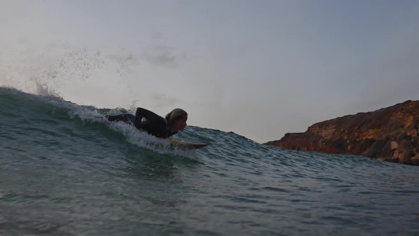 Surfer paddling on board in front of blue wave with cliffs in the background