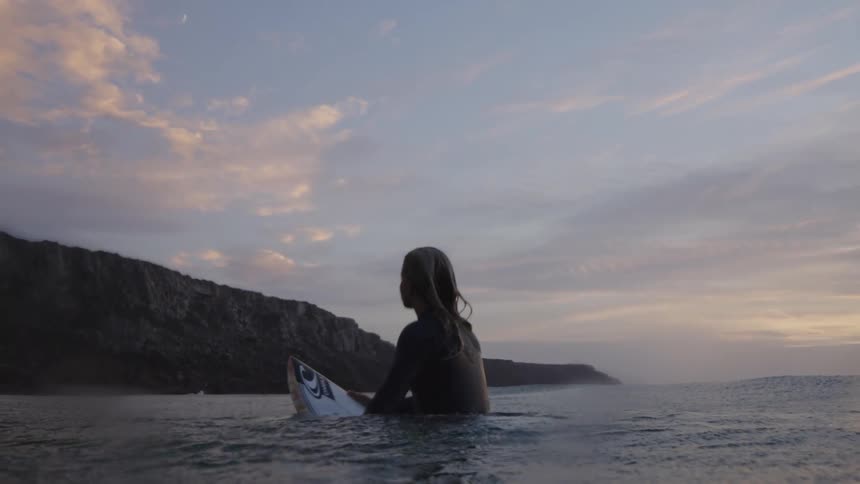 A young woman rides her surfboard in the ocean at sunset holding it steady as she glides across the glowing water framed by warm hues and gentle waves