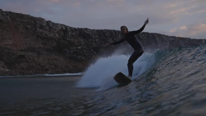 A surfer rides a large wave near a rocky cliff under an overcast sky