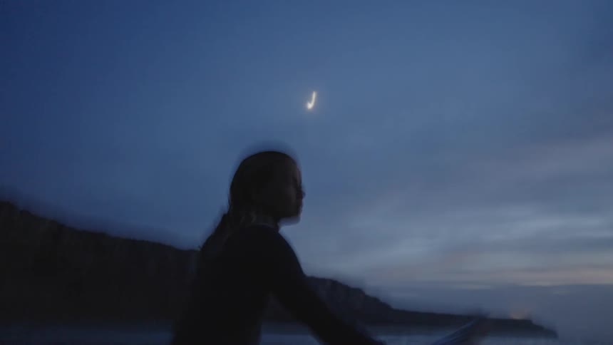 A woman is visible as a dark shape against the water sitting in a surfboard the sea surface shows gentle movement a large landmass sits in the distance all under the twilight