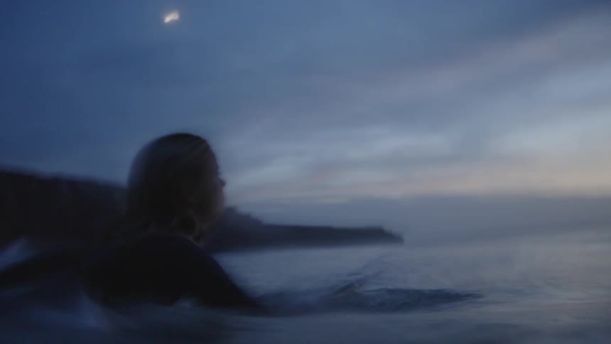 A young woman dives into the ocean with her surfboard at sunset swimming out toward the glowing horizon as golden light reflects off the waves