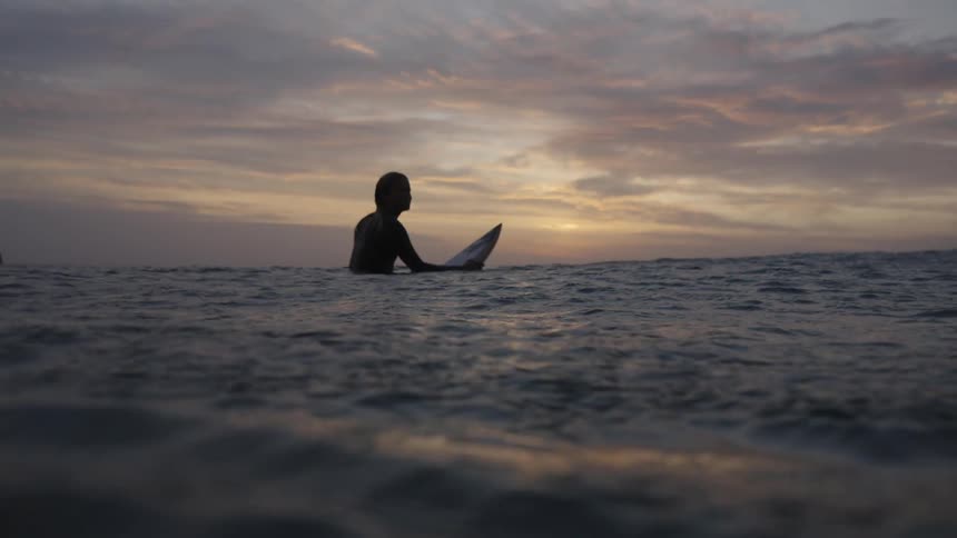 A person in a wetsuit sits waistdeep in calm dusk water silhouetted against a soft pastel sky