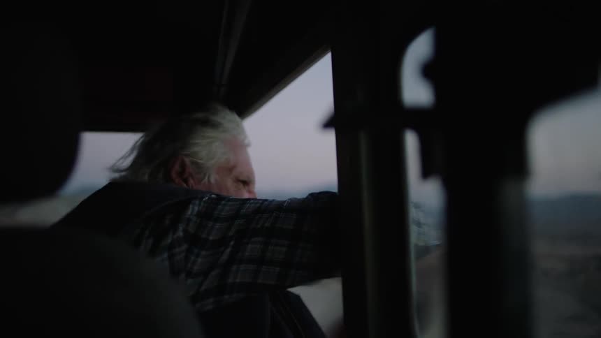 Elderly man rides in a truck waving his hat out the window
