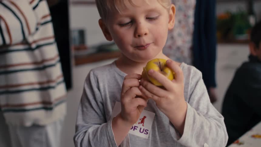 The video is a closeup of a childs hands as they hold a partially eaten yellow apple the child is wearing a gray longsleeved shirt with a sticker that reads one of us