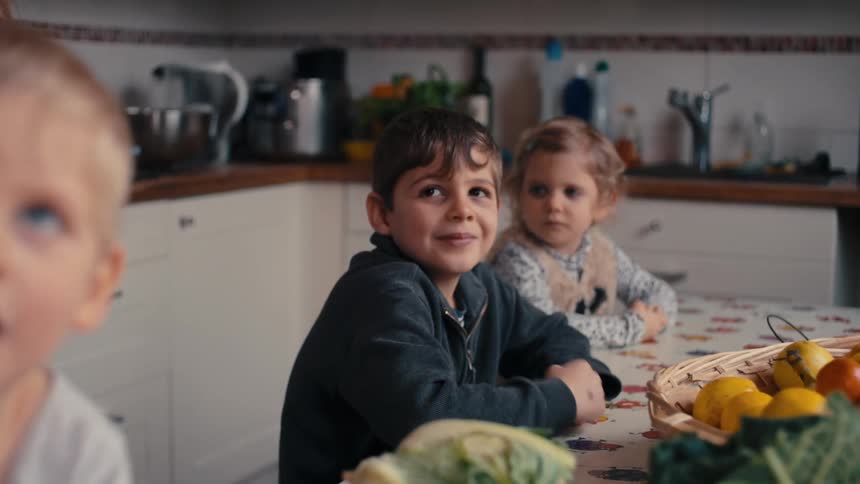 The video is a closeup shot of three young children sitting together at a table in a kitchen looking in different directions there is a large bowl of fruit and some cabbage or leafy greens on the table in front of them