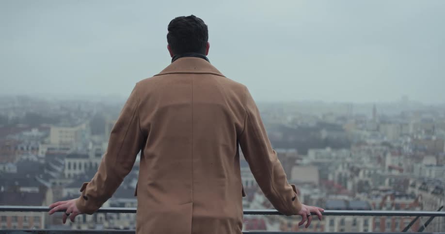 The man leans on the railing of the balcony overlooking the city