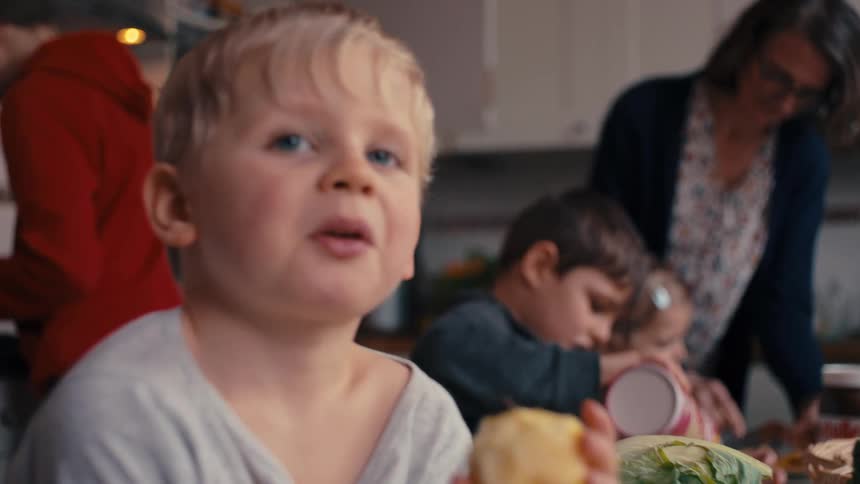 A medium view of a woman wearing glasses and a floral shirt supervising several young children eating in a bright kitchen with one blond boy smiling directly at the camera in the foreground
