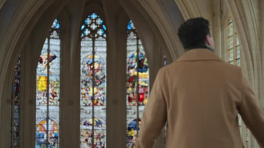 A man in a camel coat stands inside a gothic church facing tall stained glass windows