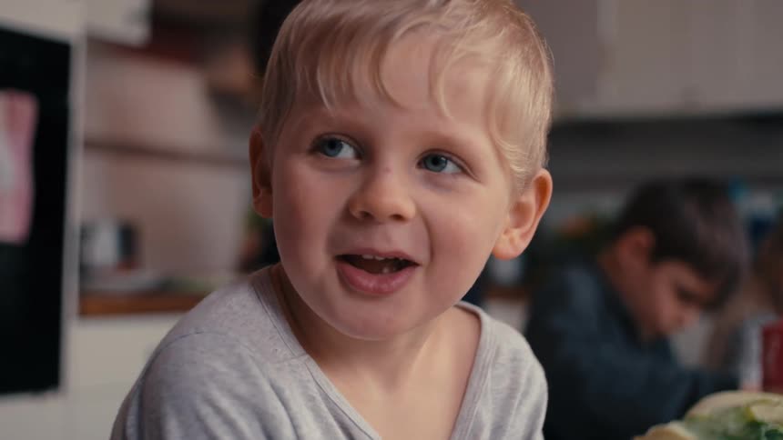 A young boy with light hair sits at an indoor surface and looks offcamera with a wideopen mouth with two other kids on the blurry background