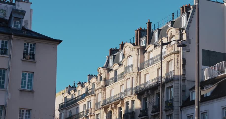 The video is an exterior shot of a row of elegant parisian apartment buildings with black wroughtiron balconies and mansard roofs a single bird is seen flying across the bright blue sky above the rooftop line