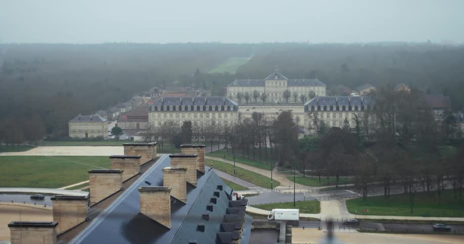 The video is an elevated wide shot of a large ornate european building set within a sprawling grassy park and surrounded by forest the sloped dark roofs and chimneys of a foreground building frame the composition obscuring part of the extens