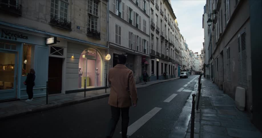 The video is an eyelevel shot of a man walking down a narrow wet street in paris the street is lined with historic lightcolored buildings featuring tall windows and commercial storefronts