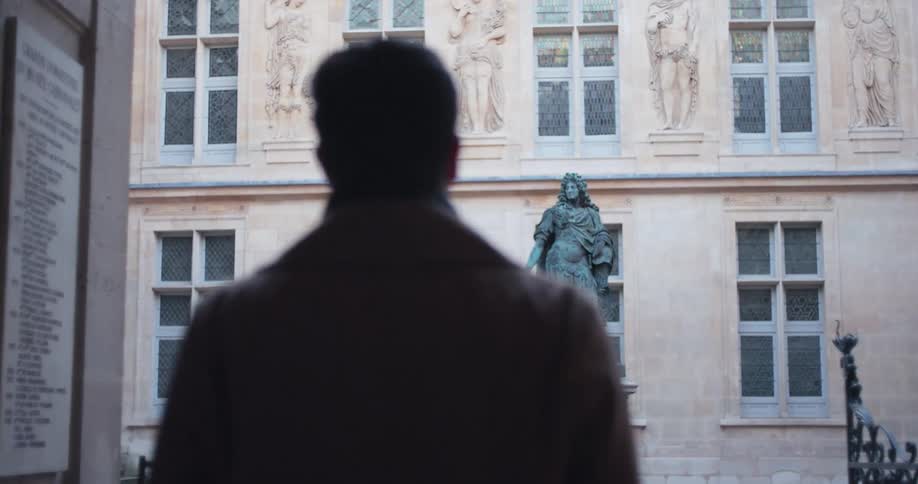 The silhouette of a man wearing a dark coat stands in a doorway looking out into a historic european courtyard as the video slowly reveals a large bronze statue standing between the stone walls and tall windows of the ornate building facade