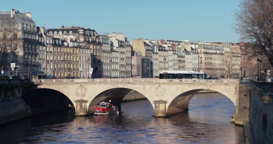 On a sunny day in paris a boat passes under an arched stone bridge on the seine river with a bus crossing above and historic buildings lining the quay