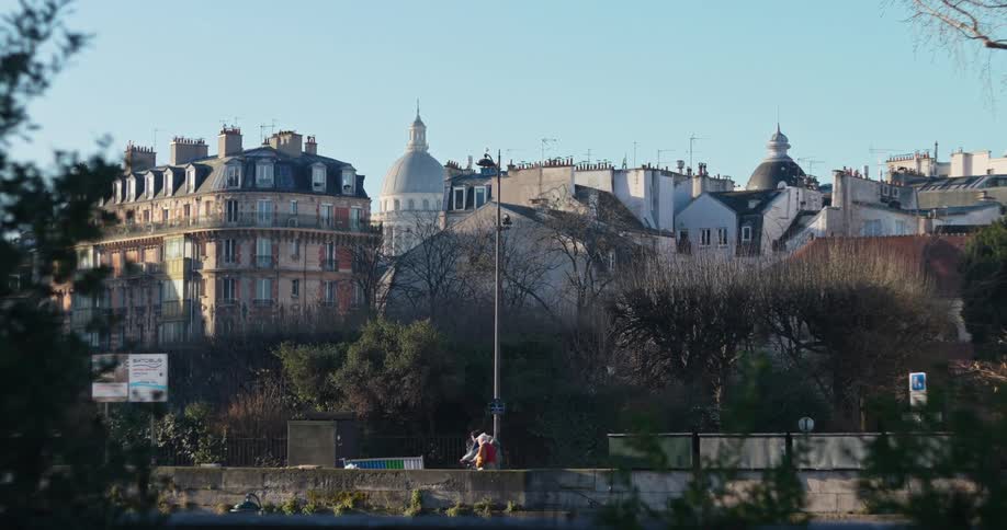 A dense collection of multistory buildings including prominent dome structures and mansard roofs positioned above a row of bare trees and a walkway where there are people passing by all under a clear sky