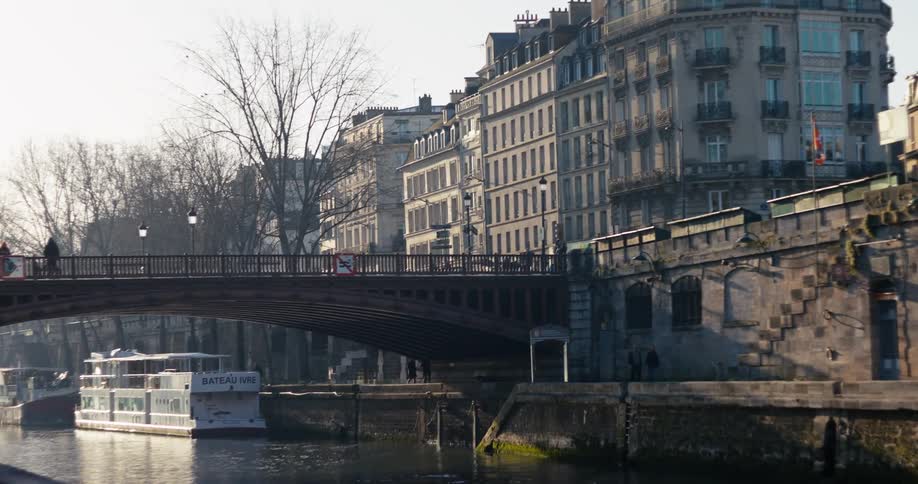 The video shows an establishing shot of a bridge crossing a river in a european city likely paris on a bright day along the banks there are houseboats or barges docked next to tall classic apartment buildings