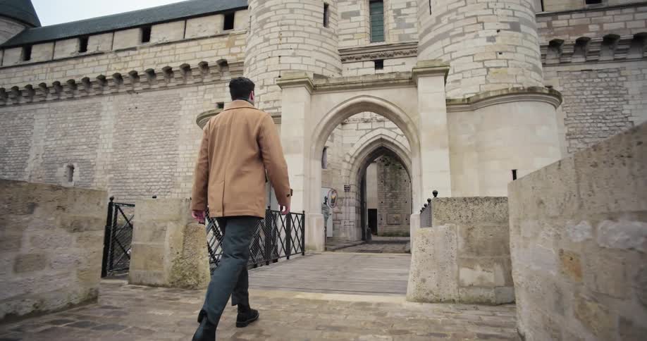 The video shows a man in a tan overcoat walking away from the camera toward the stone gate of a historic european castle or fortress the architecture features tall narrow windows and an arched entryway all constructed from lightcolored stone