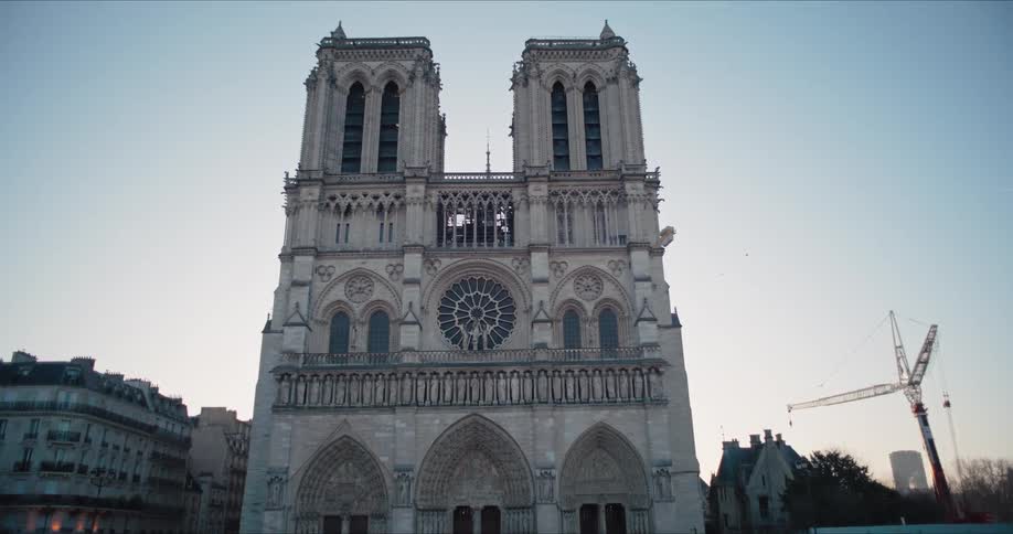 A full frontal view of a large grey stone cathedral with two symmetrical towers and a central circular window surrounded by other multistory buildings while a construction crane stands to the right under a light overcast sky and theres a crowd