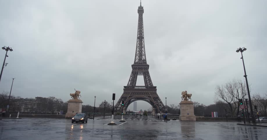 The video shows a lowangle long shot of the eiffel tower on an overcast rainy day in paris water is pooling on the street in the foreground and there are two equestrian statues on pedestals flanking the avenue that runs under the tower