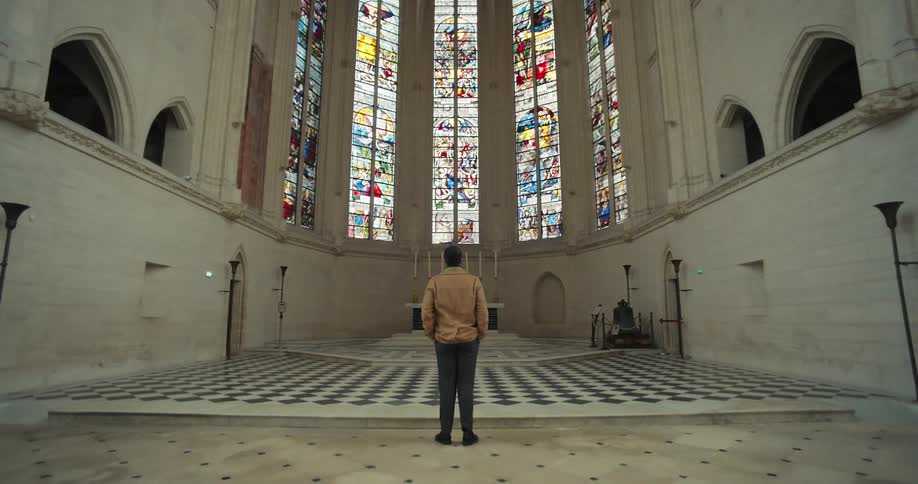 The video is an interior shot of a large cathedrallike hall with white stone walls and a vaulted ceiling a man in a tan jacket and dark pants stands with his back to the camera looking up at a series of tall colorful stainedglass windows