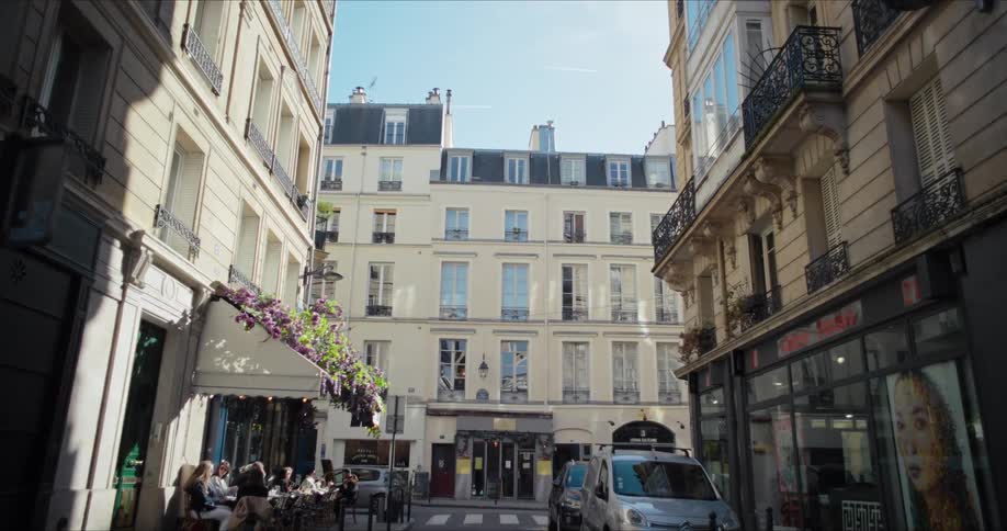 The video shows an establishing shot of a narrow street in paris with classic lightcolored apartment buildings lining both sides people are seated at an outdoor cafe on the left enjoying the sunny weather