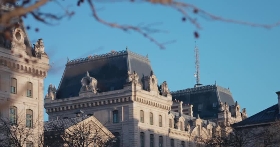 The camera starts focused on the upper portion of a large lightcolored stone building with a dark sloped roof and an antenna structure then slowly tilts upward and focuses on the bare spindly branches of a foreground tree against a bright blue sky