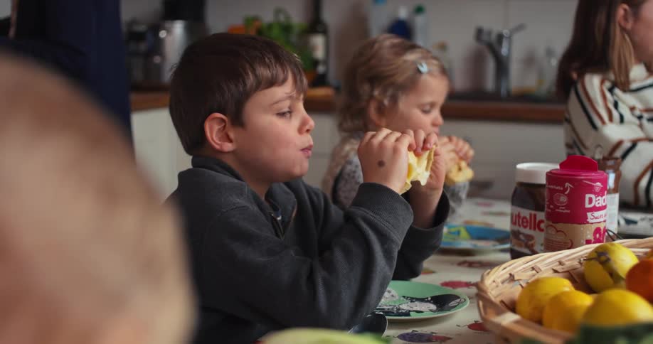 The video shows a group of children including a girl in a striped sweater and a boy in a dark tshirt sitting around a table having breakfast in the foreground another boy and a younger girl are also visible actively eating slices of bread