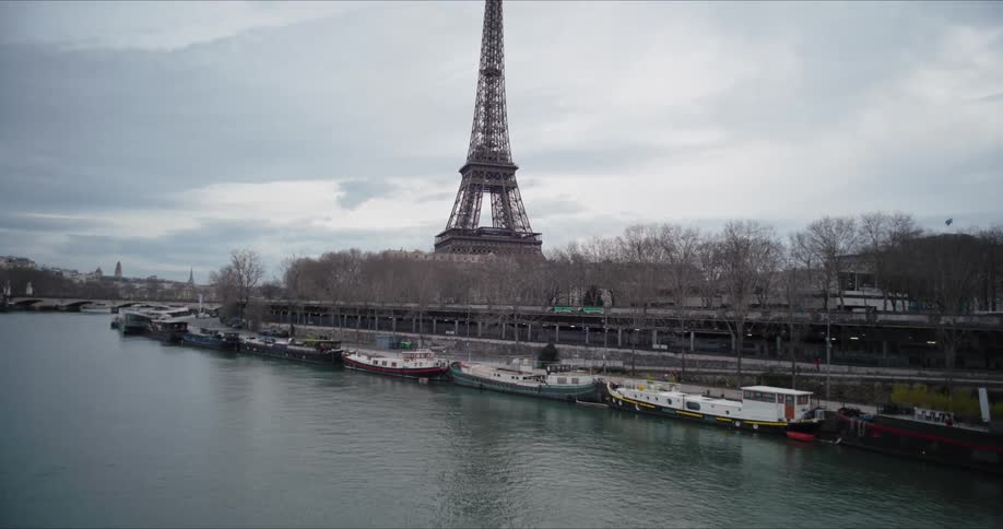 The video is a wide elevated shot of the seine river in paris with several docked barges or houseboats lining the near bank the eiffel tower dominates the center background visible above the treelined riverbank on a gray overcast day