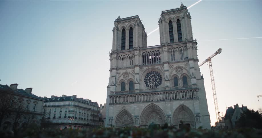 A full frontal view of a tall grey stone cathedral with two symmetrical towers and multiple arched entrances flanked by multistory residential buildings with a construction crane standing next to the structure against a pale blue sky