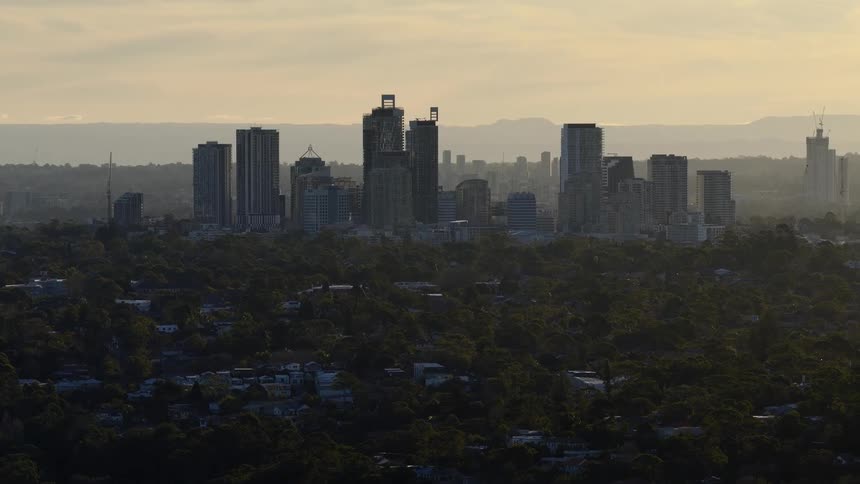 An aerial video shows a dense cluster of modern highrise buildings forming a downtown skyline rising above a vast dark expanse of low tree covered hills and suburban rooftops against a hazy pale yellow sunset sky