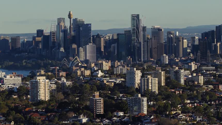 The video is a wide steady shot of a modern city skyline with many tall buildings and skyscrapers under a clear blue sky in the foreground there are lower residential buildings and a canopy of green trees that separates them from the centra