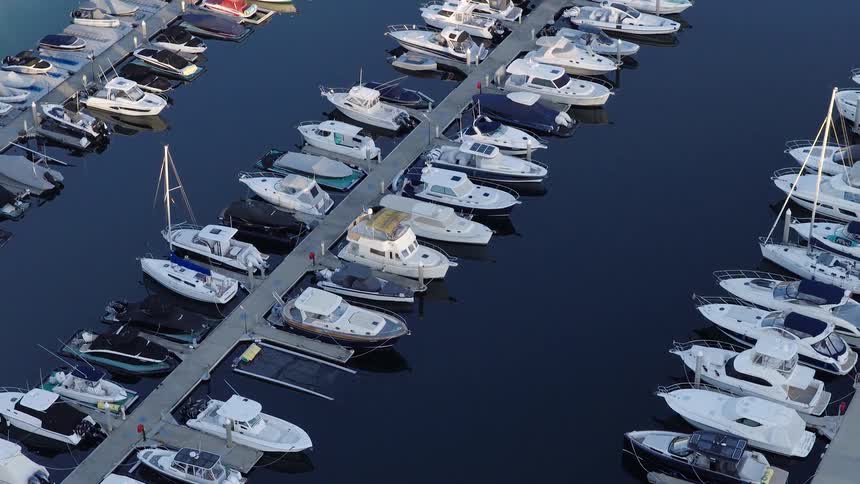 The video provides a highangle aerial view of a marina full of docked boats numerous white motorboats and small sailboats are tied neatly into their slips on the dark reflective water