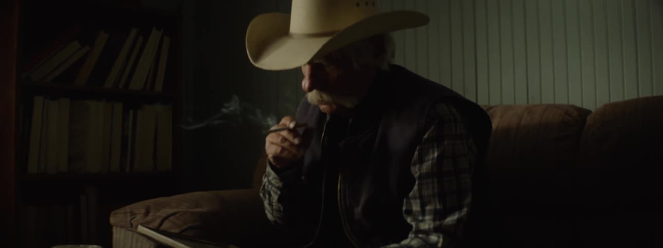 Elderly man wearing a cowboy hat smokes a cigarette while looking at a picture