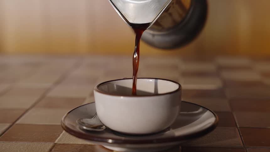Coffee being poured from a metal coffee maker into a cup on a saucer