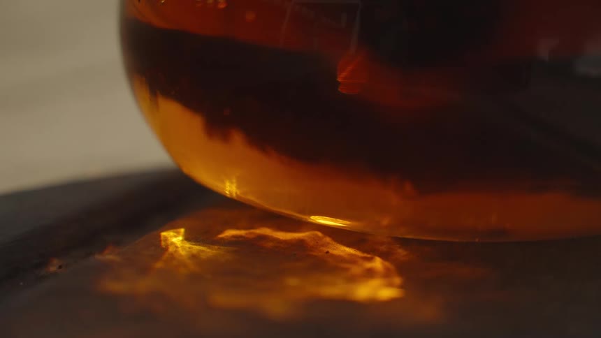 A closeup of amber liquid in a glass vessel casts warm flickering reflections onto a tabletop