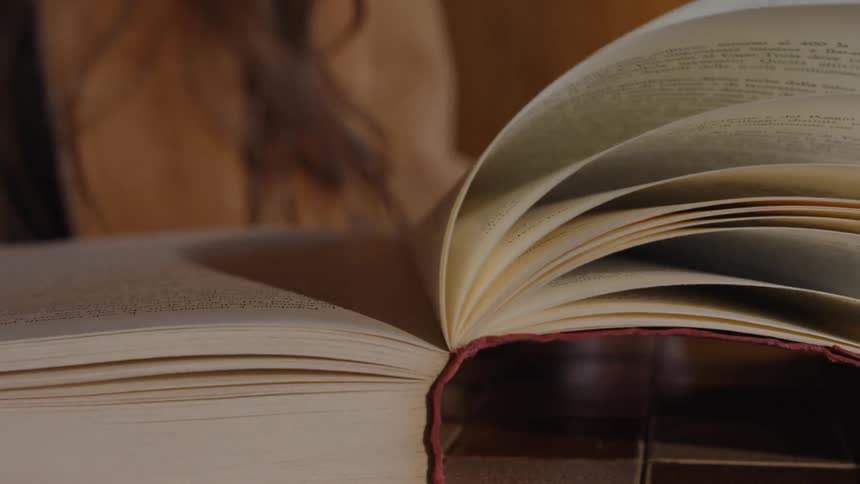 A persons fingers turn a page of a large open book with a reddish spine revealing the printed text on the exposed white paper sheets while the persons upper body remains unfocused in the background