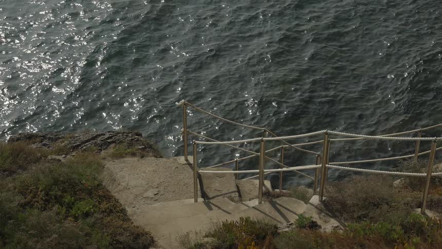 A set of steps made of lightcolored stone descends from the foreground toward a large body of dark water flanked by a rope handrail supported by metal posts and surrounded by low dry vegetation