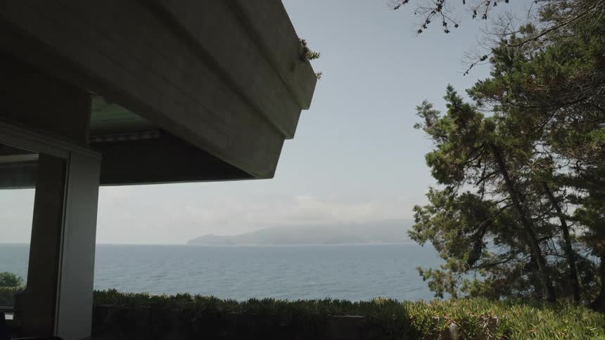 A modern concrete terrace overlooks a calm blue sea with a hazy island on the horizon framed by evergreens
