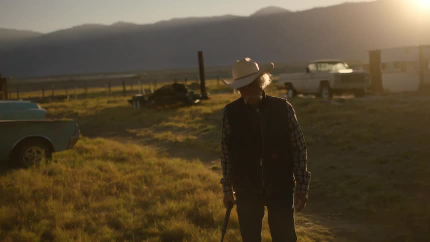 An old cowboy walks outside with a gun