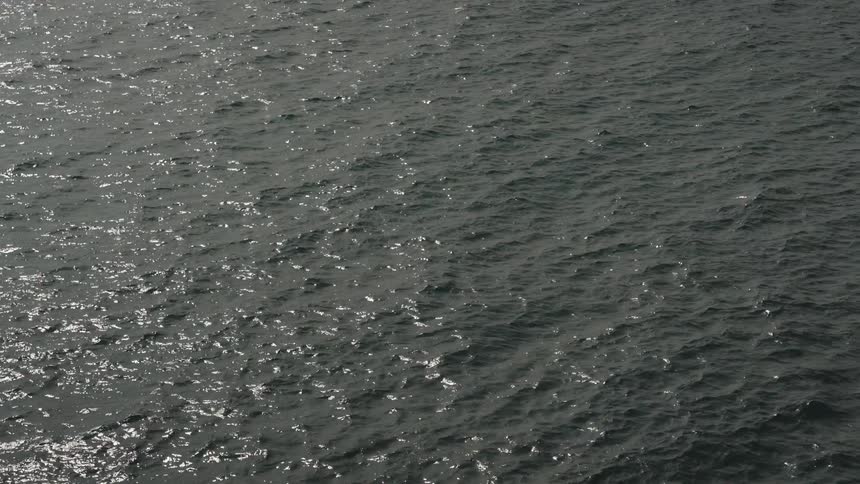 The video is an overhead shot of dark rippling water on a sunny day the surface of the water is characterized by a strong contrast between bright sunlit reflections and dark shadows