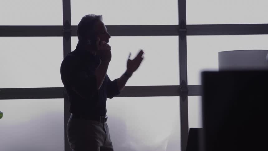 An angry man stands in silhouette on a phone call before a frosted garage door window one hand raised as he gestures