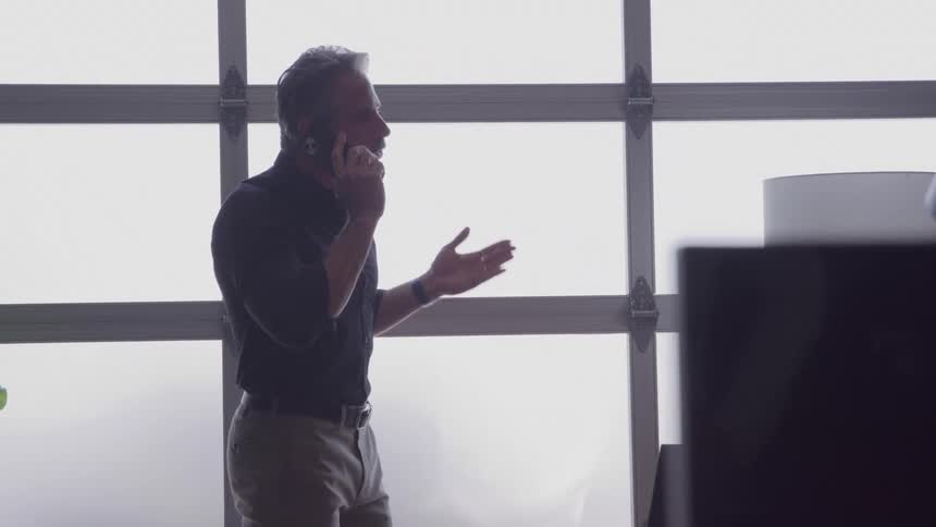 The video features a middleaged man in a black shirt and tan pants standing indoors near a large frosted window wall he is actively speaking on a mobile phone while gesturing with his free hand