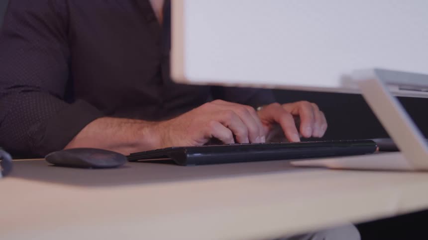 A man types on a keyboard at a desk beside a monitor and a laptop
