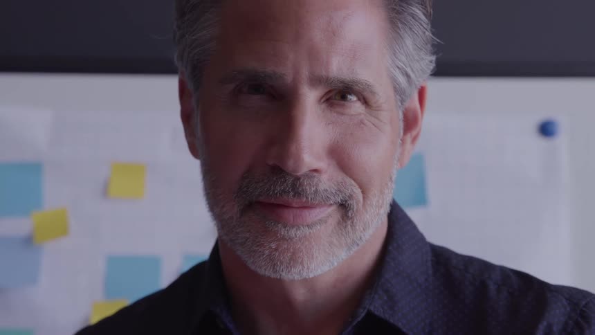 Older man standing near a whiteboard with post it notes on it smiles