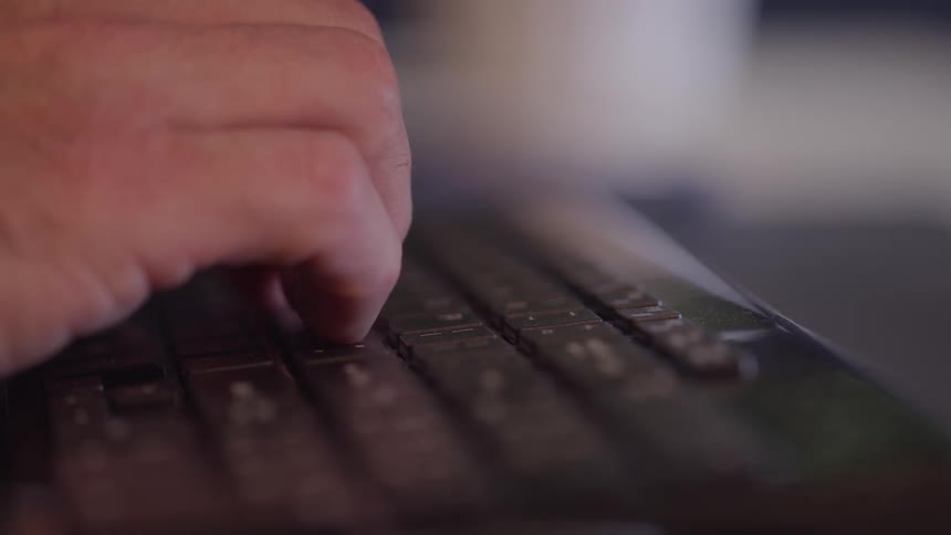 A close view video shows a persons fingers rapidly and firmly typing on the dark keys of a laptop or computer keyboard in a dimly lit interior setting