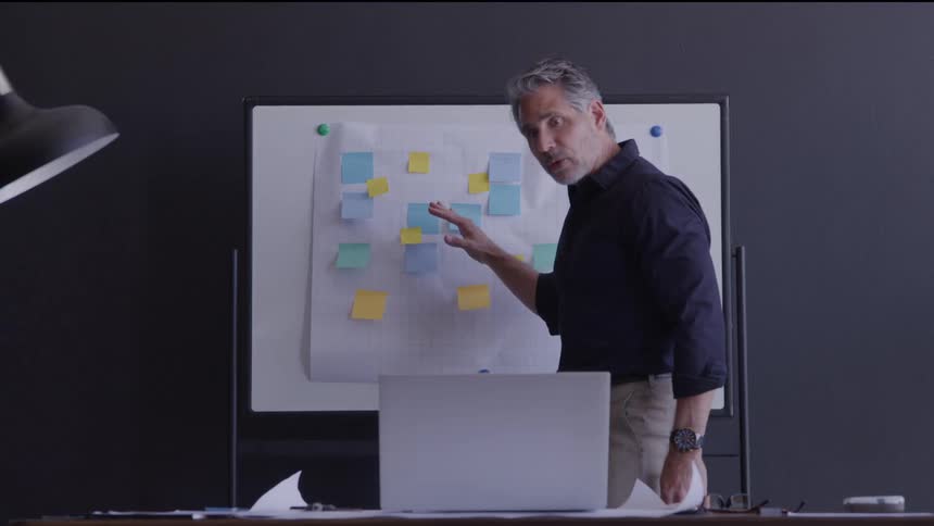 A middle aged man with gray hair wearing a dark shirt and khaki pants stands over a silver laptop at a desk looking up to gesture and speak towards it as a white board covered in blue and yellow sticky notes stands behind him