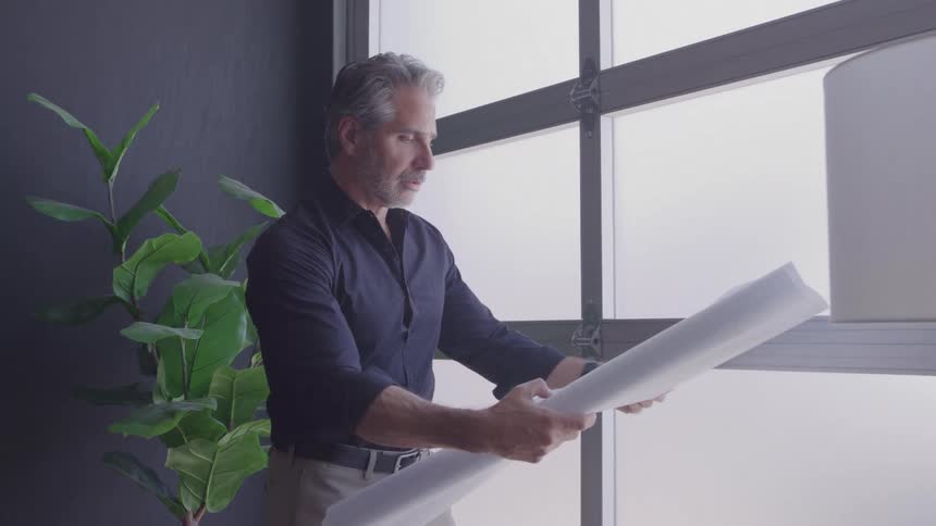 A man with gray hair in a dark shirt studies a rolled blueprint by a frosted door beside a tall indoor plant