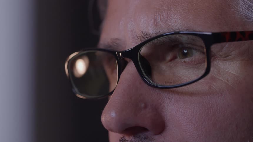 A macro video close view focuses on the face of an older man with gray hair showing a reflection of a bright screen in his dark framed glasses as he blinks while staring intently forward in a dimly lit setting