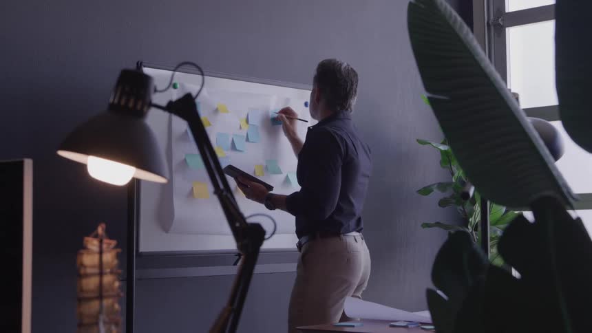 A man stands in an indoor area and faces a white board covered with small colored papers holding a dark digital tablet in one hand and writing on the board with a pen in the other hand while a bright lamp illuminates the foreground