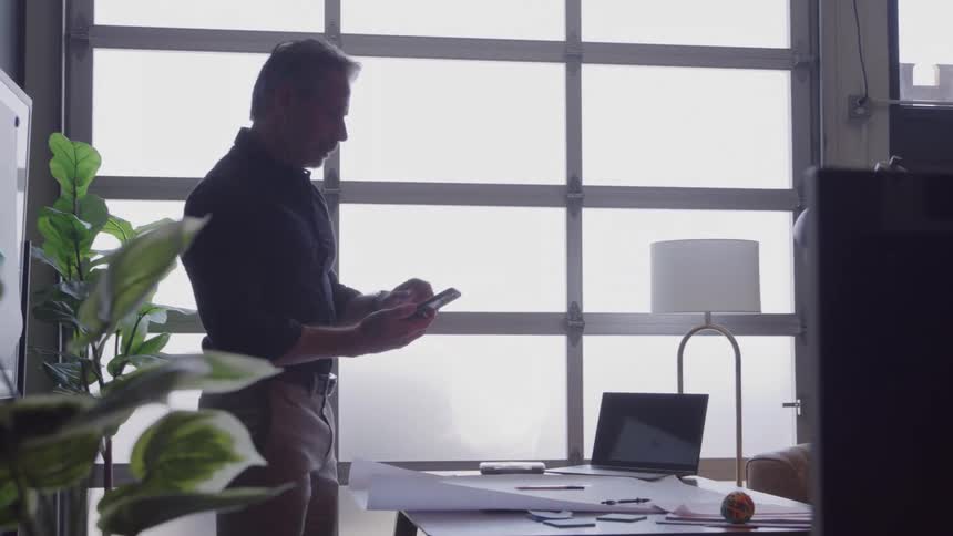 A professional older man in a dark shirt and khaki pants works at a desk with blueprints and a laptop before stopping to answer his mobile phone near a large bright window in a modern office space